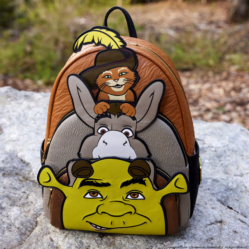 Image of the Loungefly Shrek, Donkey, & Puss in Boots Exclusive Triple Pocket Mini Backpack sitting on a gray rock outside with dirt and grass in the distance behind it 